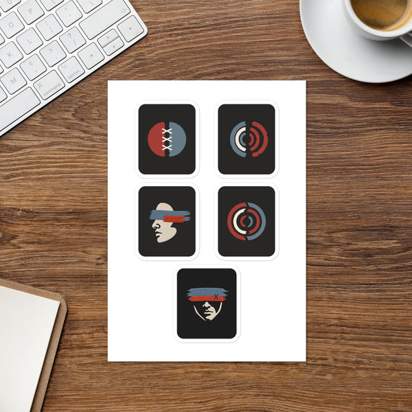 Five-design Witness sticker sheet on a desk beside a keyboard and notebook; both face designs are visible.